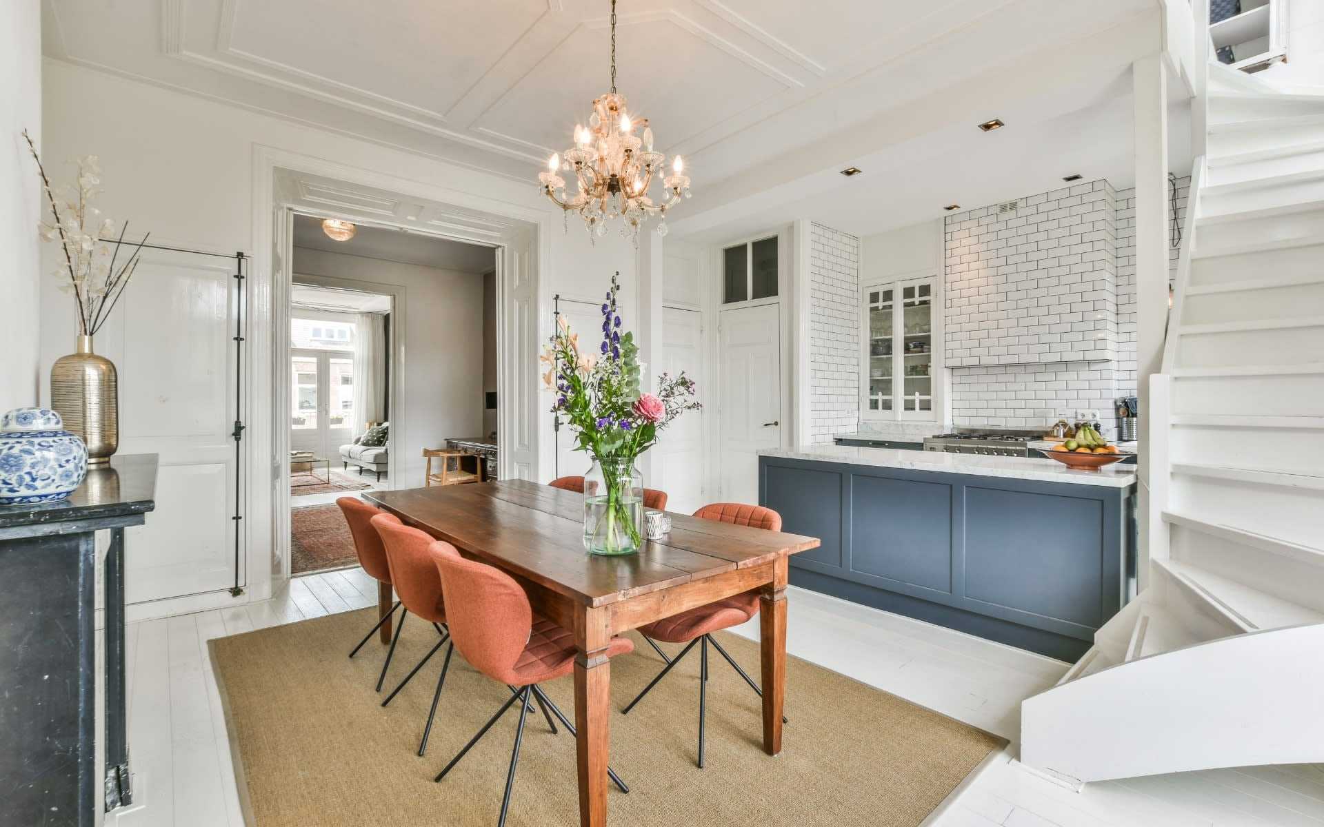 A dining room with a wooden table, orange chairs, and a chandelier, leading to a blue kitchen and a living room.