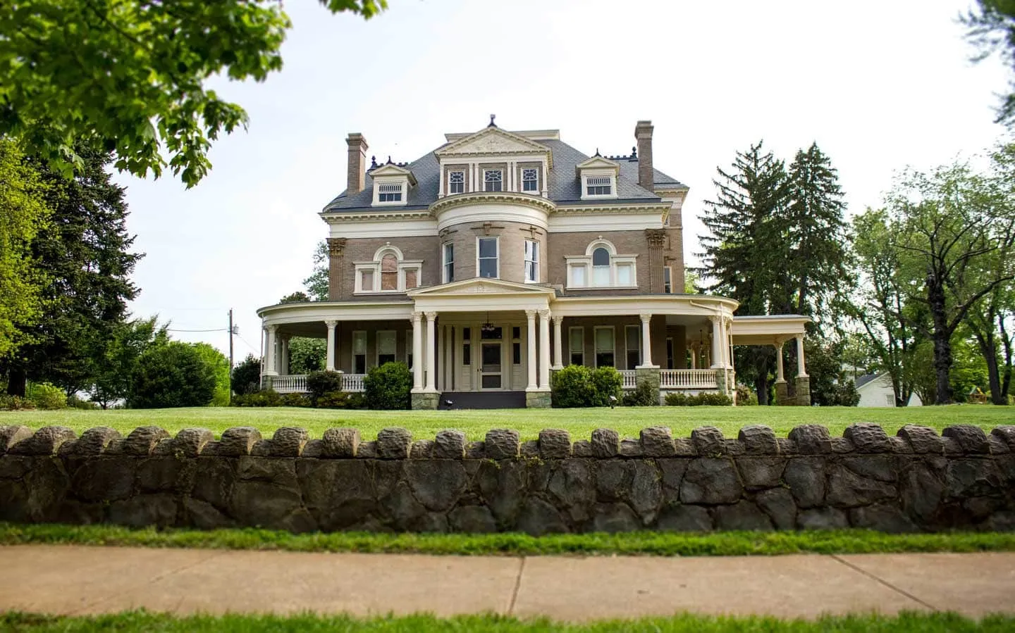 Large historic home with a wraparound porch above a stone wall